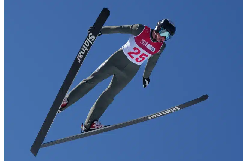 An Olympic ski jumper in mid-air against a clear blue sky, wearing a helmet, goggles, and a red bib labeled 25. The scene conveys focus and exhilaration — but now rumors of hyaluronic acid.