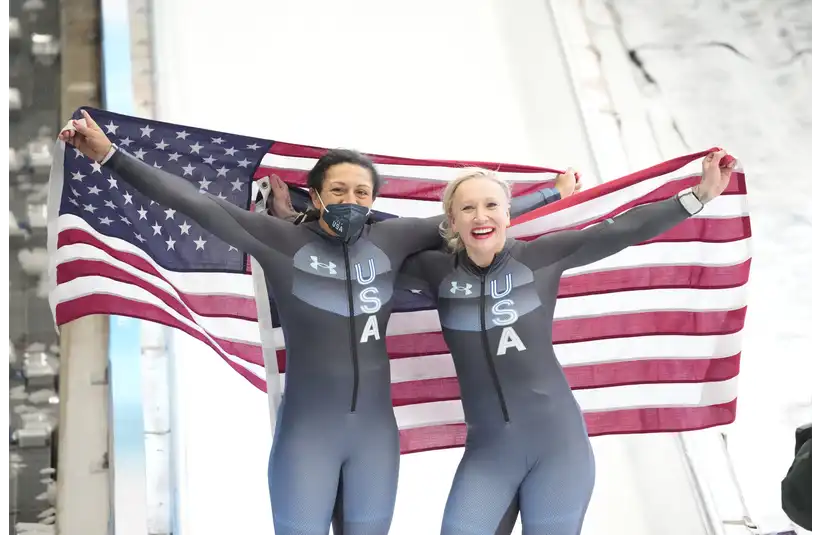Two Olympic athletes in USA uniforms celebrate joyfully, holding a large American flag behind them on a snowy track. One wears a mask, both smiling broadly.