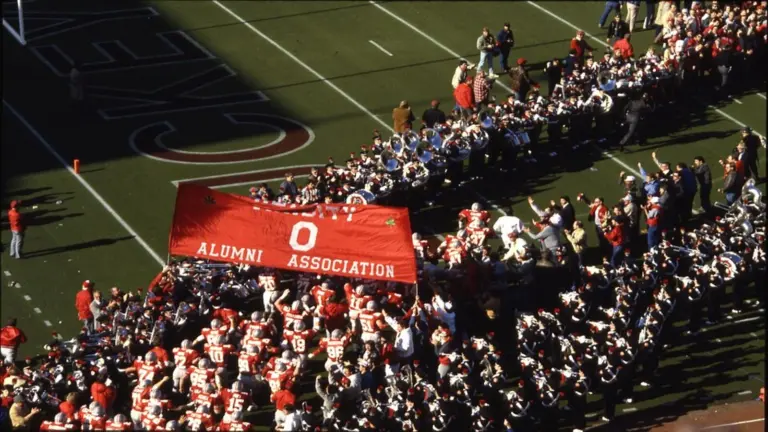 A view of the 1987 Ohio State Buckeyes football team as they run out onto the field in Ohio Stadium. Members of the Varsity O Alumni Association hold a banner for the players to run under and, along with the marching band, they form two lines for the players to run between.